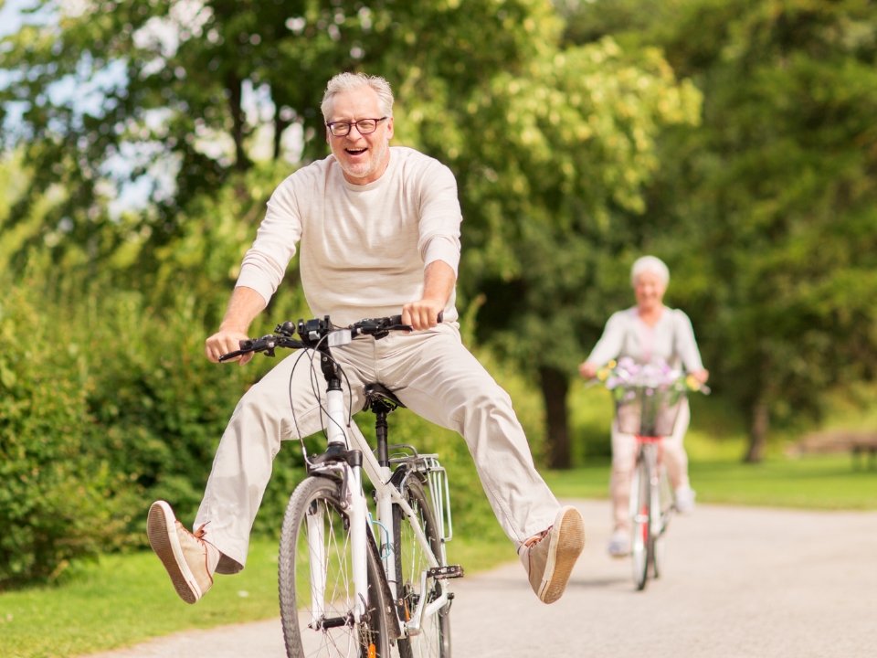 Older man joyfully riding a bike with his legs out, partner riding behind
